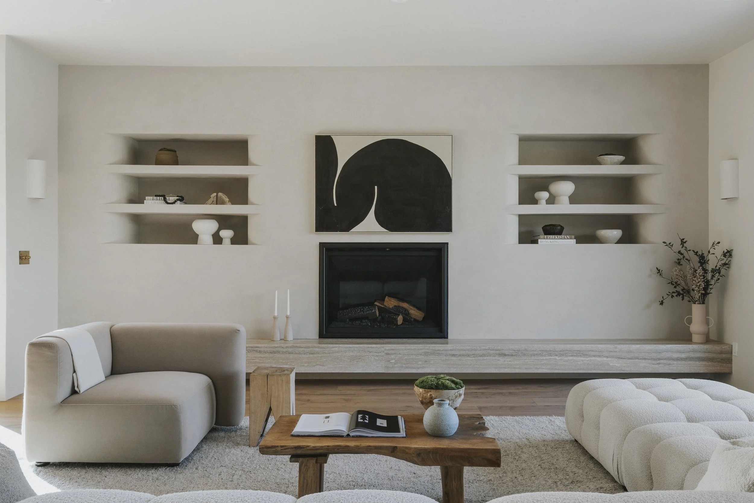 A living room with a sculptural black artwork above a fireplace, built-in plaster shelving, and soft neutral seating.